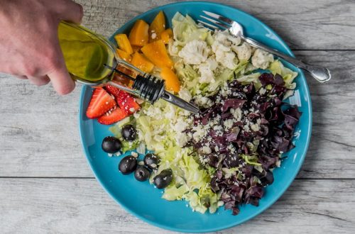 A vibrant vegetable salad with olives, feta, and strawberries being drizzled with olive oil.